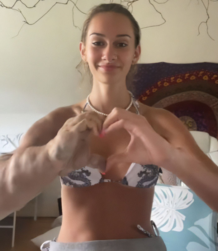 Smiling woman with one larger arm forming a heart with both hands in a relaxed indoor setting.