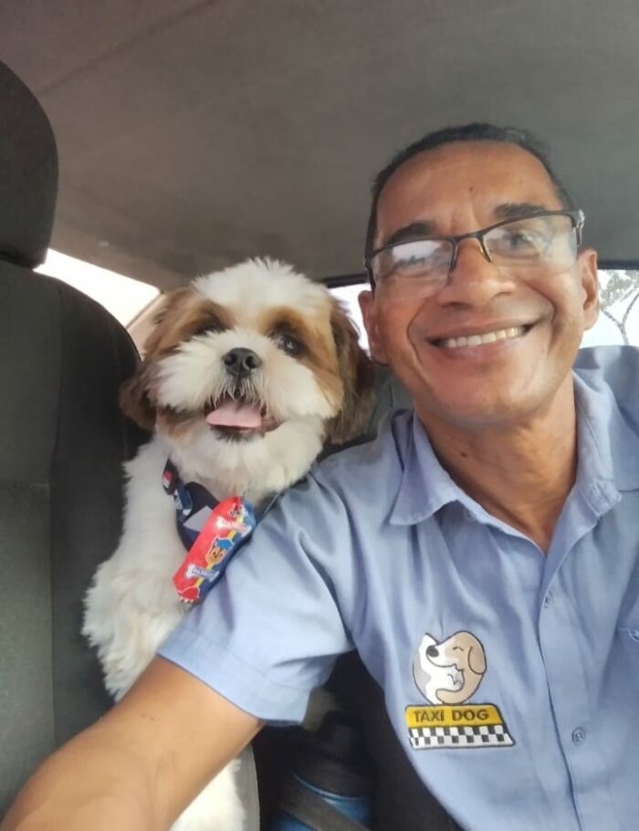 Man smiling in pet taxi selfie with fluffy dog wearing a colorful bandana in the passenger seat.