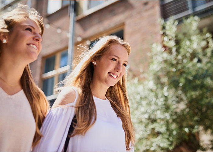 Two women walking outdoors on a sunny day, smiling, with a focus on their joyful expressions.