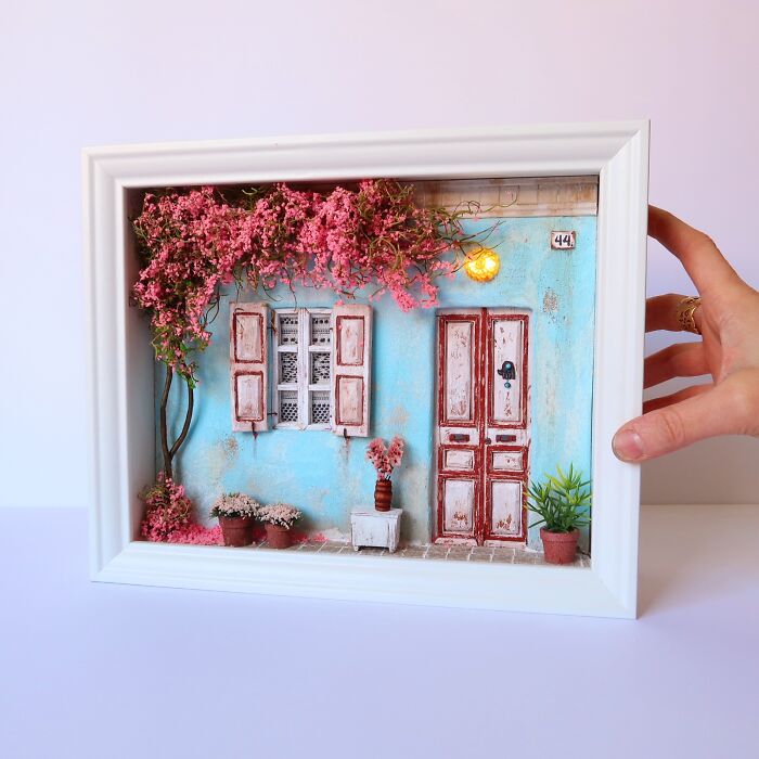 Miniature art by Tamar Cohen showcasing a tiny, detailed facade with pink flowers, held in a white frame.
