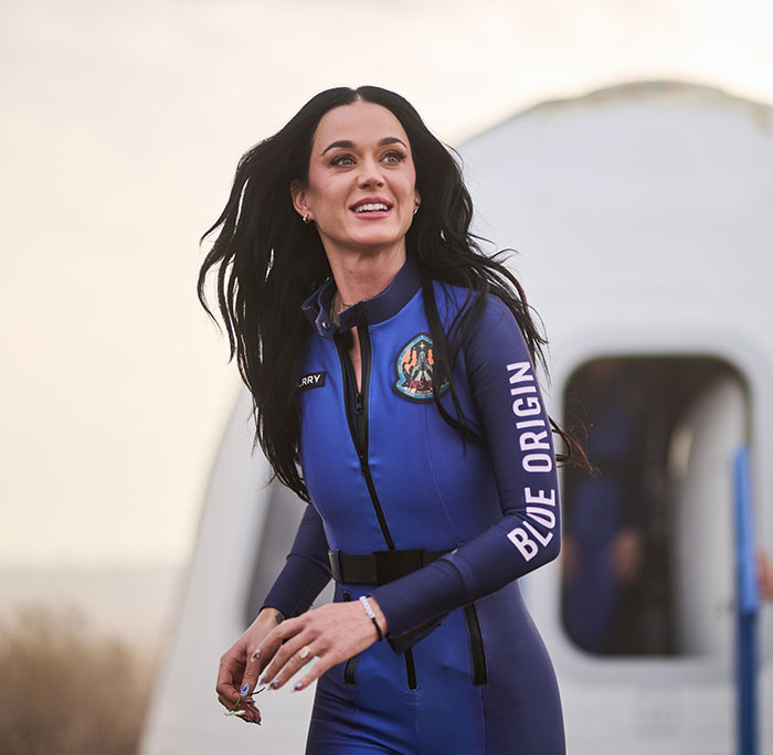 Person in Blue Origin spacesuit, outdoors, with futuristic setting in background.