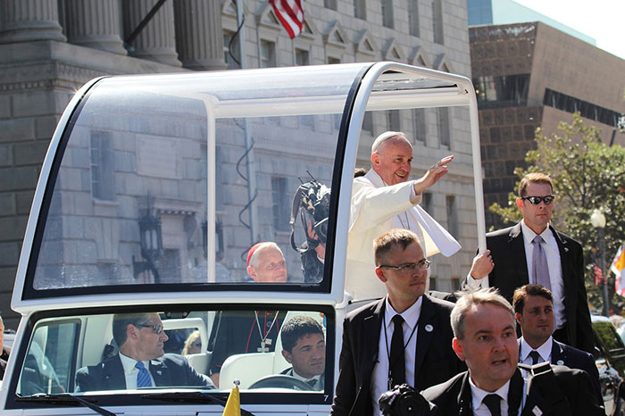 Pope Francis standing in an open popemobile, waving to a crowd, escorted by security personnel.