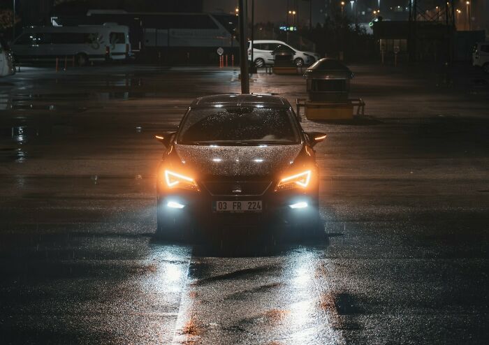 Car with headlights on at night in a wet parking lot, evoking a sense of potential danger.