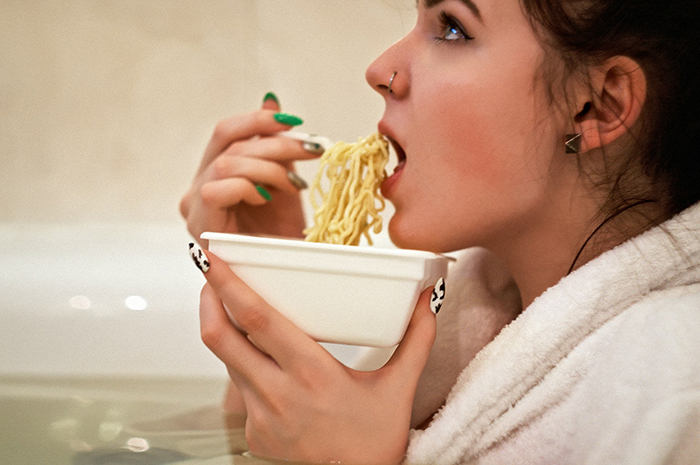 Woman eating noodles in a bathtub, highlighting a gluten-free meal setting.
