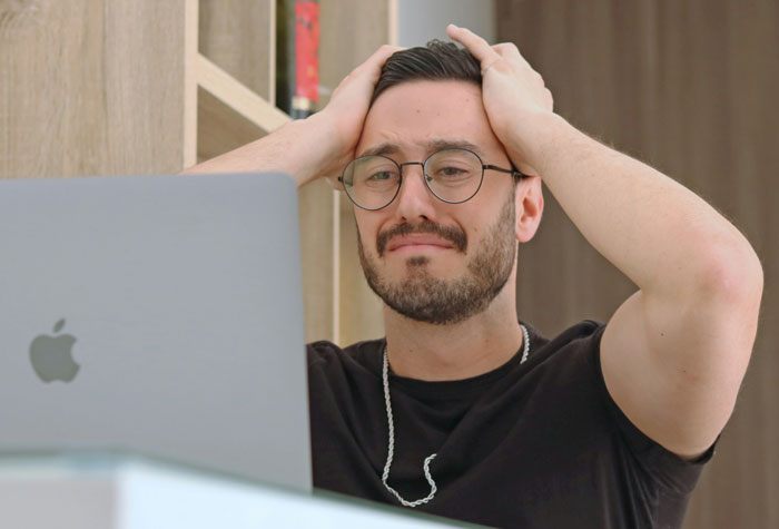 Man in glasses looks upset at laptop, hands on head, questioning work balance.