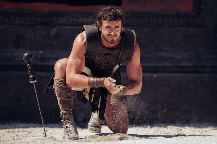 A man in gladiator armor, kneeling in a sandy arena, representing beloved stars in a heroic scene.