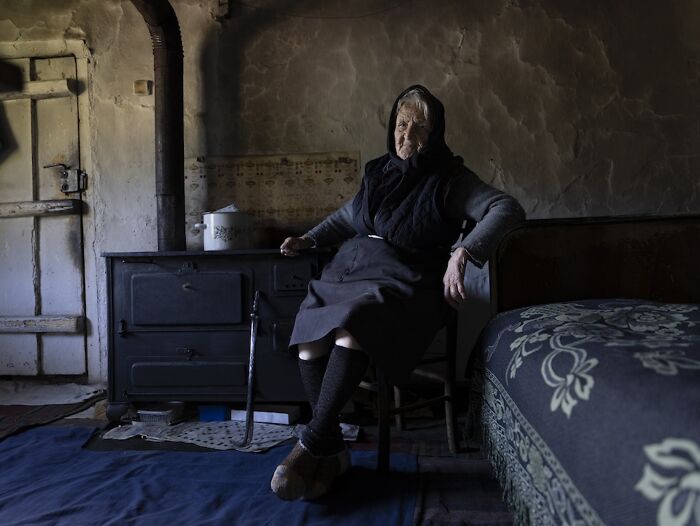 An elderly woman sitting in a dimly lit rustic room, conveying the vulnerability of aging women.