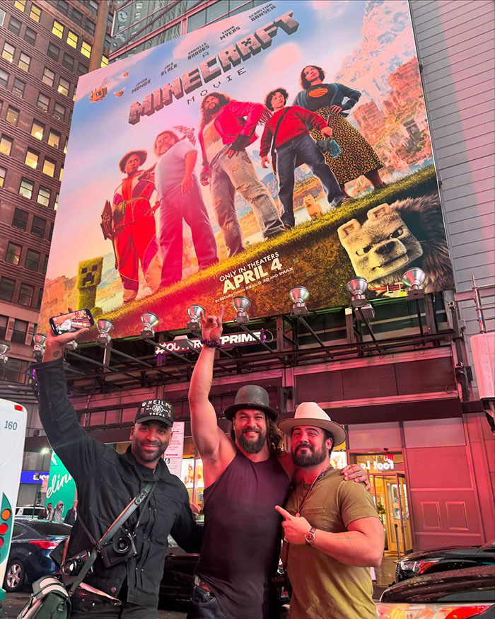 Three people pose under a large "Minecraft" movie poster in a city setting at night.