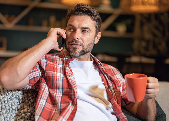 Man in a plaid shirt on a phone call, drinking from a red mug, related to Duke Dennis hug discussions.