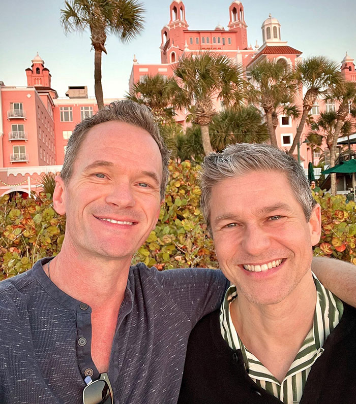 Two smiling men posing in front of a pink building with palm trees, enjoying a sunny day.