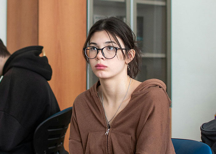 Student in a classroom setting, wearing glasses and a brown hoodie, looking thoughtful.