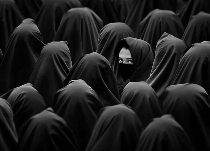 Black and white street photo of a crowd with one person wearing a mask standing out among others in dark clothing.
