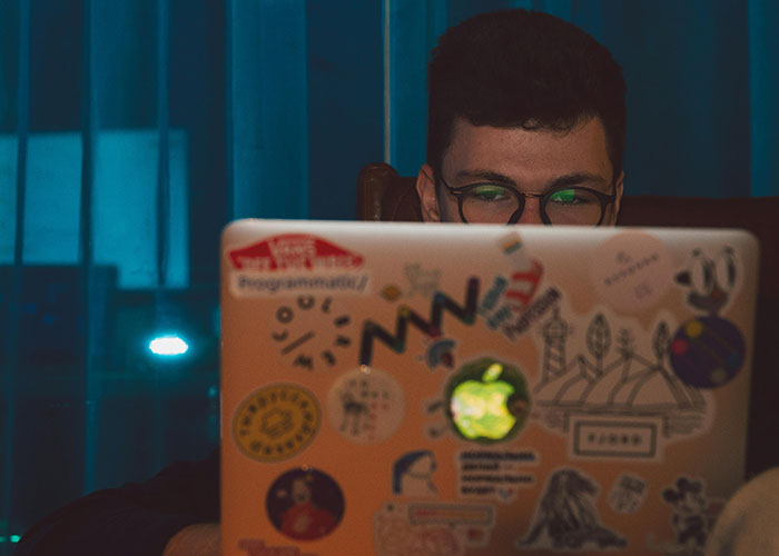 Person with glasses working on a laptop covered in stickers in a dimly lit room, reflecting heartbroken and cheating themes.