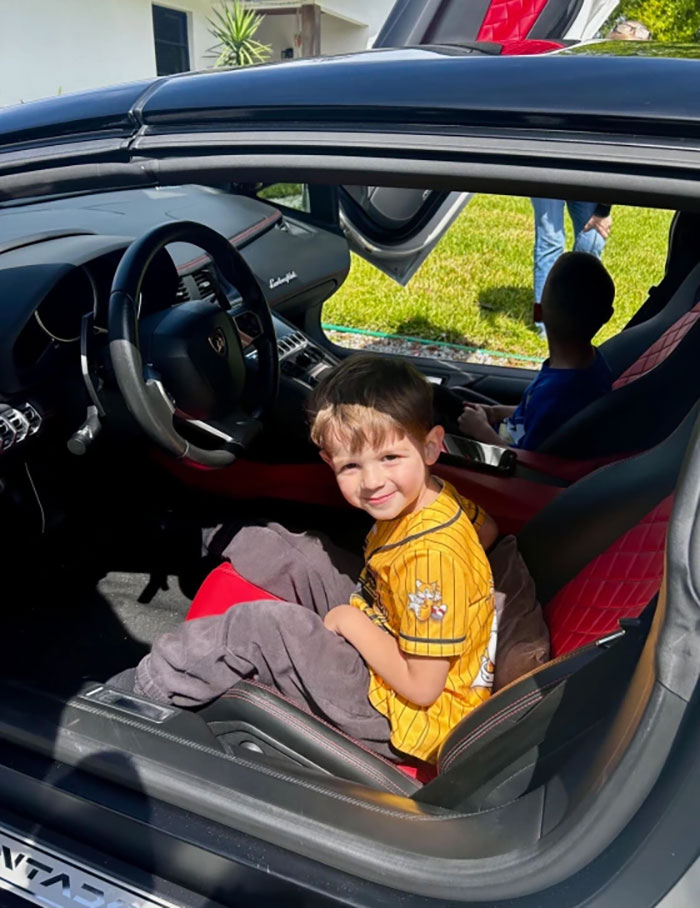 Child smiling in a Lamborghini sports car with open scissor doors, enjoying a birthday surprise from local car enthusiasts.