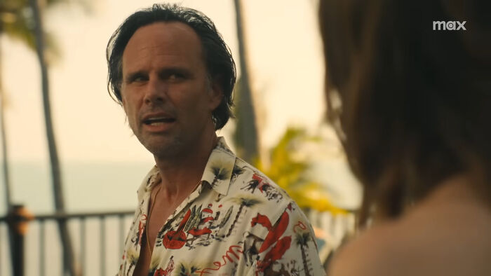 Walton Goggins outdoors in a tropical shirt, speaking with someone, palm trees in the background.