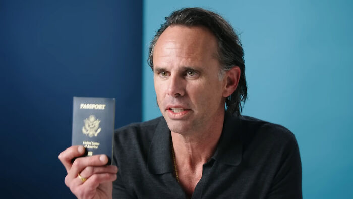 Actor holding a passport, discussing essential personal items, in a blue room.