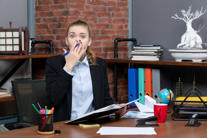 A surprised woman in an office, reacting to work drama, sits at a desk with open book and colorful stationary.