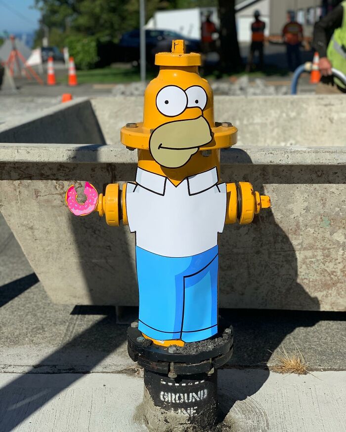 Simpsons paper cutout of Homer Simpson on fire hydrant, holding a donut, blending with real-world scene.
