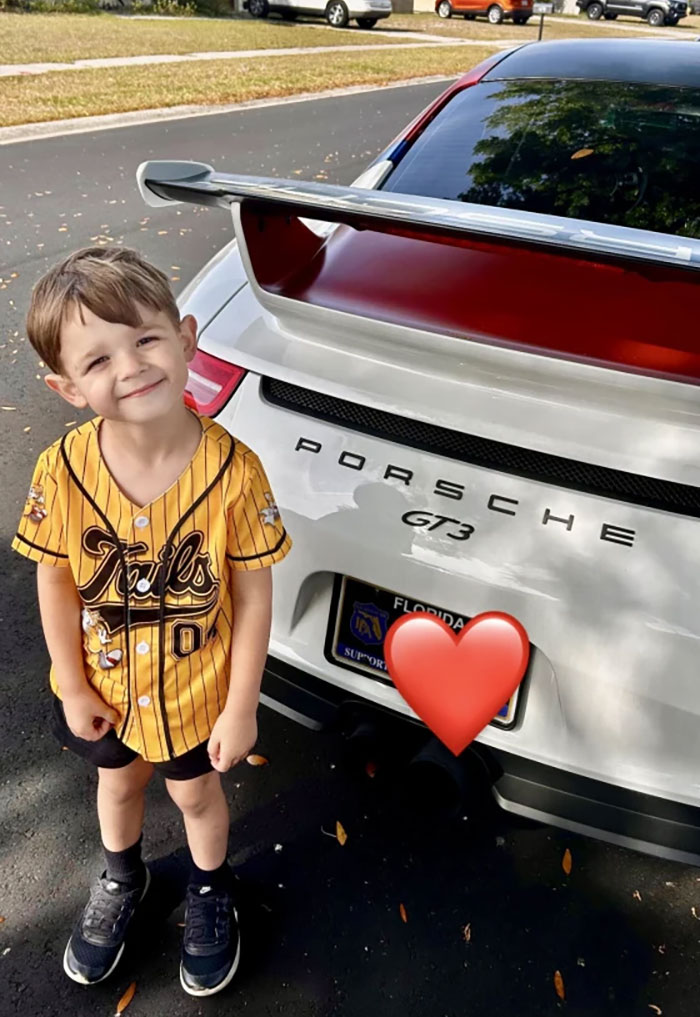 Smiling child standing next to a sports car with a large spoiler, enjoying a birthday surprise.
