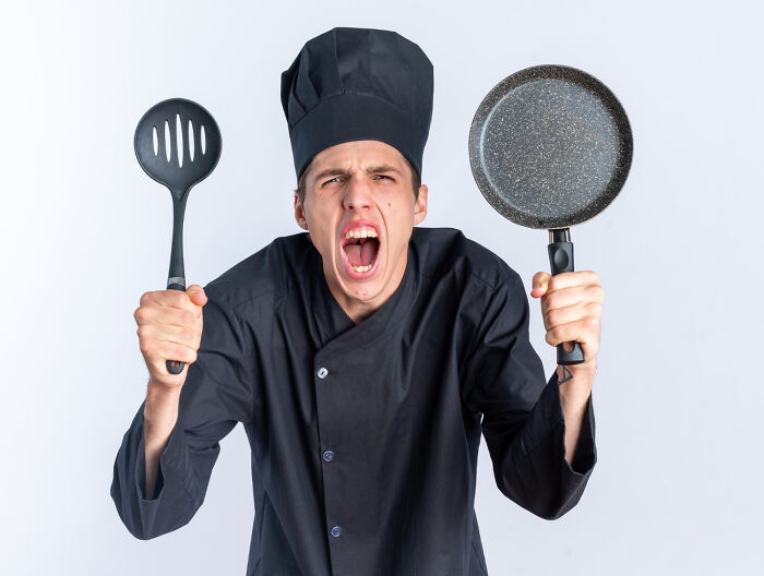 Chef in black uniform, holding a spatula and a pan, looking dramatic.