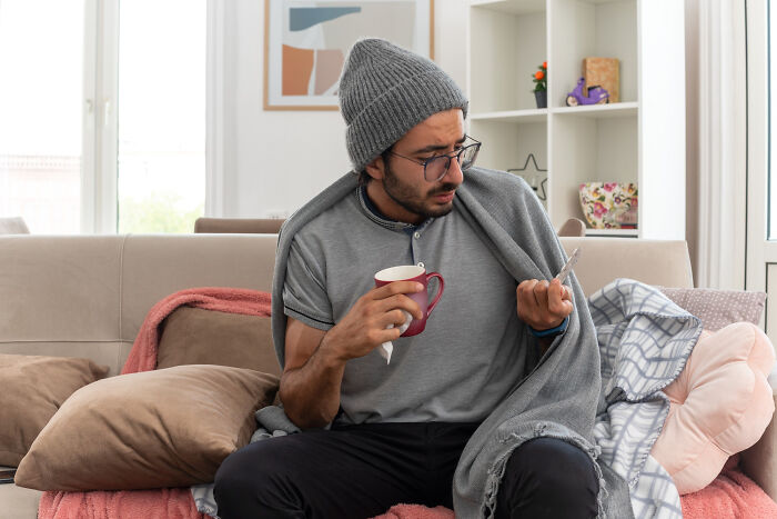 Man in gray shirt and beanie, holding a mug, looks confused at phone.