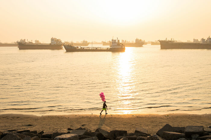 Person with pink balloons walking on a beach at sunset, ships on the horizon. World Food Photography Awards 2025 shortlist.