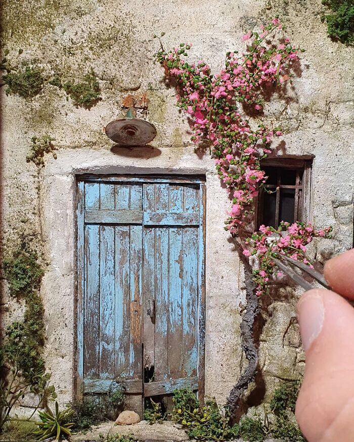 Miniature scene with a blue weathered door and pink flowers, crafted by hand to resemble a secret world.