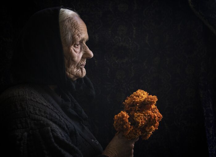 Elderly woman in profile, wearing a black hood, holding orange flowers, capturing vulnerability of aging.
