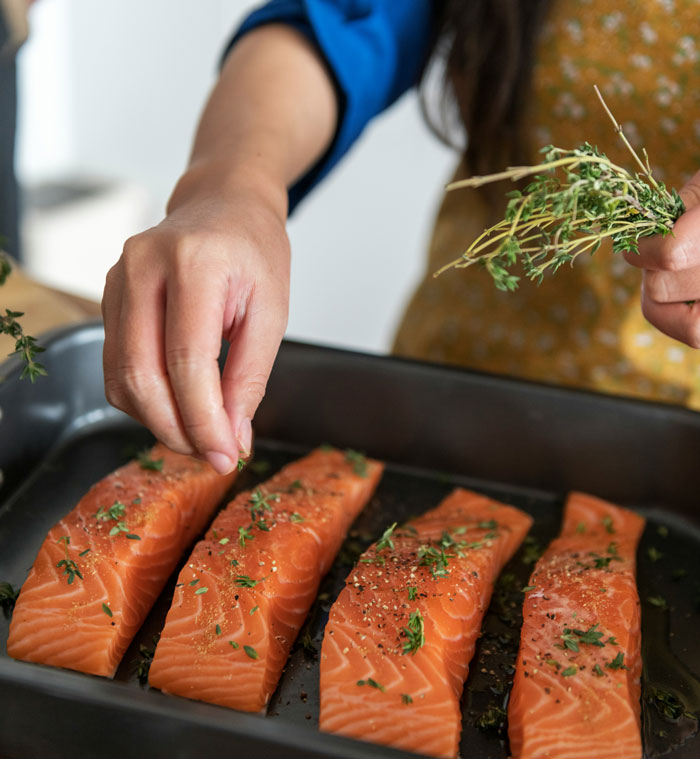 Person seasoning salmon fillets with herbs in a kitchen.