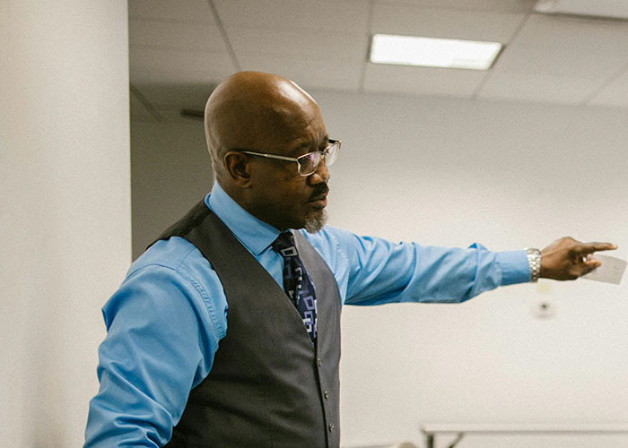 Teacher in a classroom setting, wearing glasses and a vest, gesturing while giving a lecture.