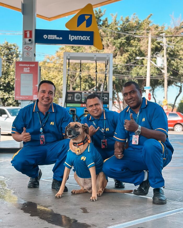 Three gas station workers and a dog wearing matching blue uniforms posing together at a gas station, highlighting the hero dog.