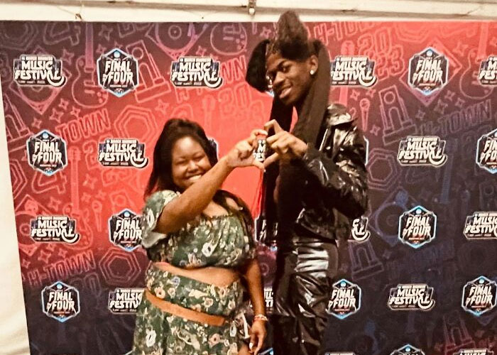 Person and celebrity wearing black outfit making heart shape with hands, smiling at music festival backdrop.