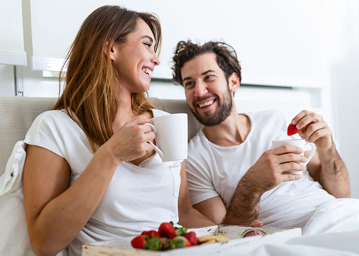 Happy couple enjoying coffee and strawberries in bed, sharing a relaxed moment and discussing marriage cheat codes.