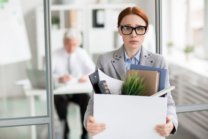 Woman in office holding a box of items, looking worried, embodying work drama scene.