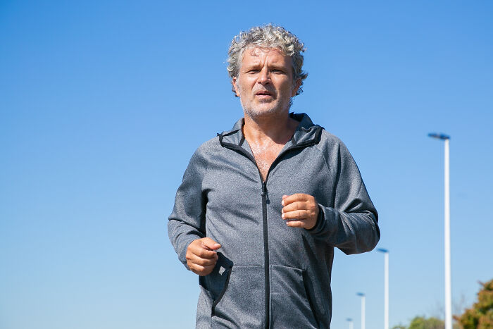 Older man jogging outdoors, embracing the realities of aging, in a gray tracksuit under a clear blue sky.