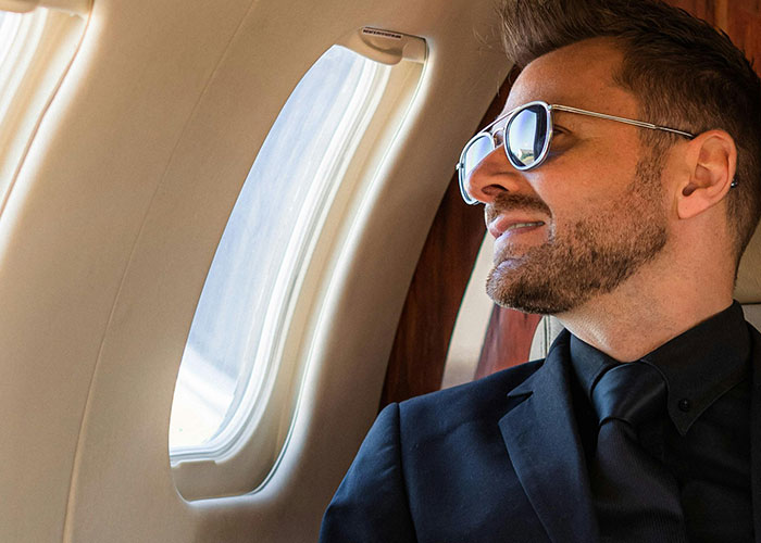 Man in sunglasses and a suit gazing out an airplane window, conveying a sense of luxury and surprise on a plane.