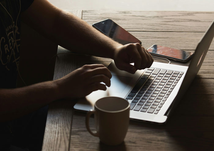 Person typing on a laptop at a wooden table with coffee and smartphones, focusing on SEO deliverables and work hacks.