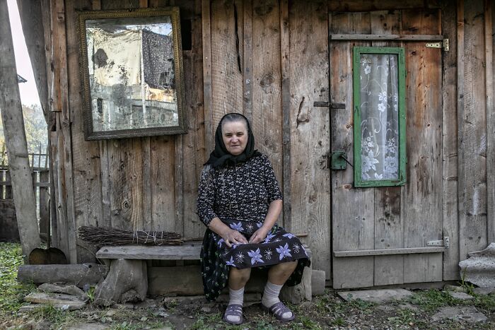 Aging woman seated outside a rustic wooden house, reflecting vulnerability and strength.