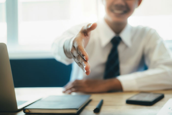 Smiling person in a suit offering a handshake across a desk, symbolizing job hunting opportunities.