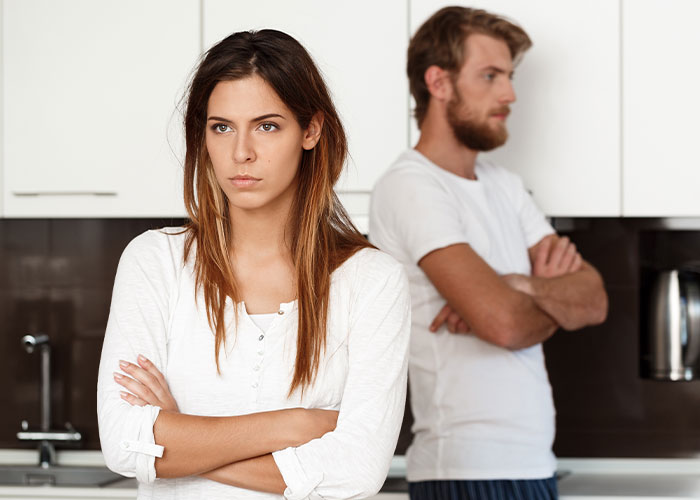 A couple stands back-to-back in a kitchen, arms crossed, displaying tension.
