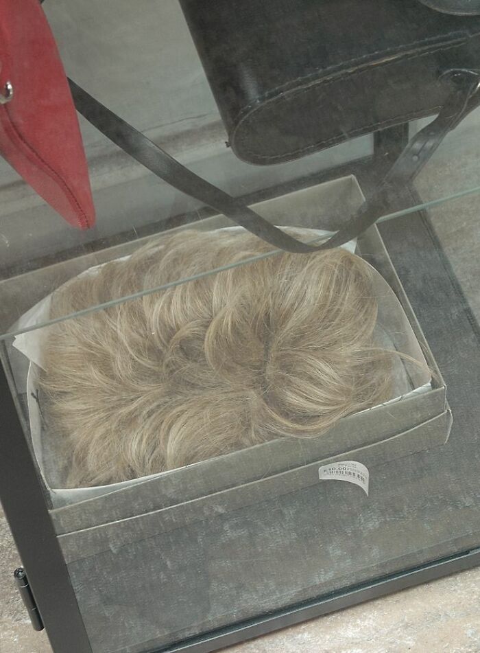 Blonde wig displayed in a secondhand shop, stored in a transparent box for viewing.