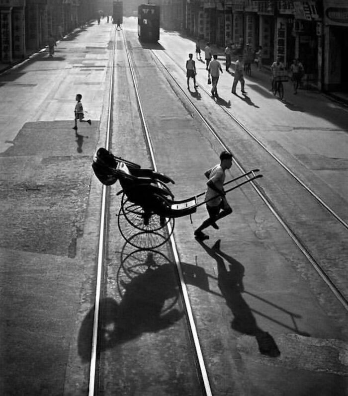 Black and white street photo showing a man pulling a rickshaw with pedestrians walking along tram tracks.