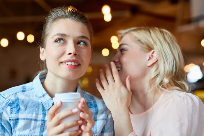 Woman whispering secrets about girlfriends to a friend holding a coffee cup.
