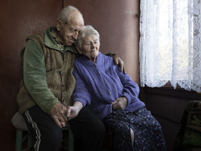 Elderly couple sitting together, conveying vulnerability of aging women, holding hands in a warm embrace near a window.