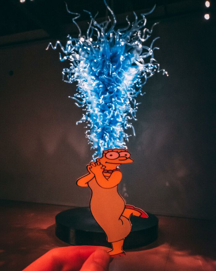 Simpsons paper cutout in front of a vibrant blue glass sculpture, creating a lively real-world scene.