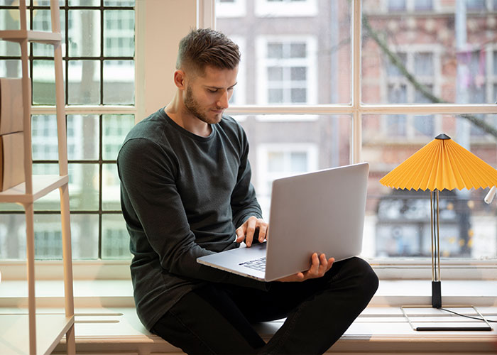 Man sitting by a window using a laptop, illustrating unrealistically serene work settings often portrayed in movies.