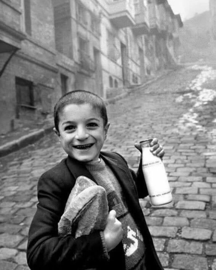 Black and white street photo of a smiling boy holding bread and a milk bottle on a cobblestone street.