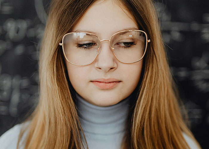 Young woman with glasses stands in front of a chalkboard, contemplating.