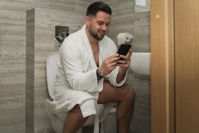 Man in a bathroom, wearing a white robe, sits on a toilet using a smartphone, illustrating dating and relationship humor.