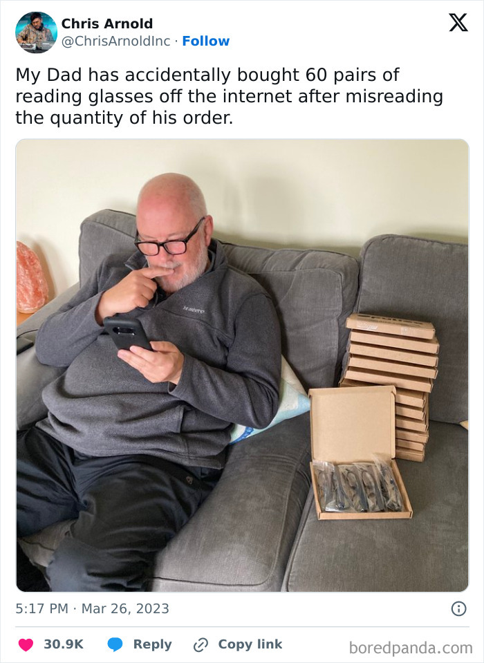Man on couch with boxes of reading glasses, looking at phone, having a worse day after accidentally ordering 60 pairs online.